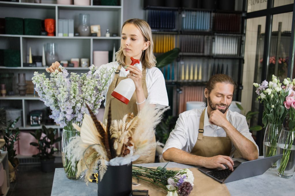 Small family business concept. Two workers caucasian businessman professional designer, florist decorator using laptop at floral store checking inventory of shop and contractor girl waters flowers.